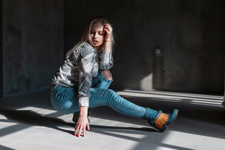 European fashionable young woman model in stylish jeans in green fashionable cowboy boots in a trendy shirt poses sitting in a room with sunlight. Beautiful blond girl model. Modern youth style.の写真素材