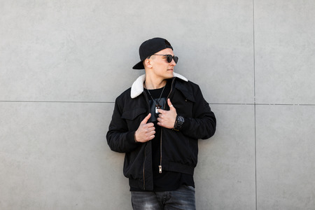 Young stylish hipster man in a baseball black cap in black sunglasses in a trendy black jacket with...