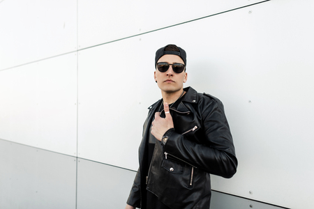 Attractive stylish young hipster man in stylish leather jacket in trendy black baseball cap in trendy dark sunglasses poses near modern white building in the city. American guy on the street. Retro.の写真素材