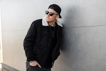 Modern stylish young hipster man in a stylish black jacket in a fashionable cap in trendy dark sunglasses in vintage gray jeans posing near a gray concrete wall. American guy. Stylish menswear.の写真素材