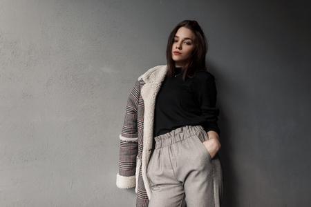Sexy beautiful young brunette woman in an elegant checkered jacket in a trendy black shirt in stylish gray pants standing near a vintage wall indoors. Pretty girl brunette fashion model. Retro style.の写真素材