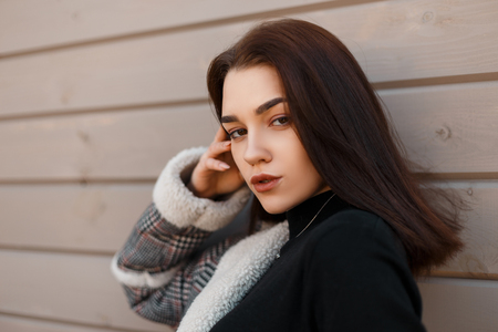 Attractive young brunette woman in a stylish gray checkered jacket in a black fashionable shirt near the modern wooden vintage wall outdoors. Beautiful sexy girl model. Portrait of a woman's face.の写真素材