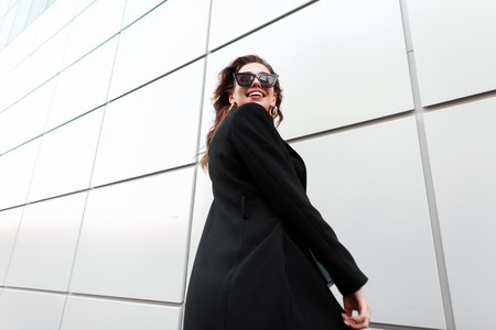 Positive pretty young stylish hipster woman in trendy dark sunglasses in an elegant black long coat is spinning and smiling near a modern building in the city. Cheerful girl walks in the spring day.の写真素材