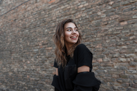 Joyful pretty young woman in a trendy coat in a black stylish T-shirt with a positive beautiful smile is standing outdoors near a vintage brick wall. Cute happy girl enjoys a walk on a spring day.の写真素材