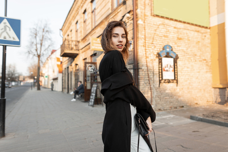 Elegant modern young woman in a long vintage coat with a black leather handbag in a trendy white jeans in a T-shirt walking down the street on a sunny day. Beautiful girl model relaxes at sunset.の写真素材