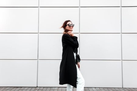 Pretty joyful young hipster woman in a vintage black long coat in white jeans with a stylish leather handbag in dark sunglasses is posing near a modern white wall. Happy cute girl on a walk.の写真素材