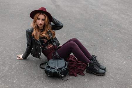 Beautiful young woman in a chic hat in a leather jacket in purple pants in stylish boots with a fashionable backpack relax sitting on the asphalt on a spring day. Attractive red-haired girl resting.の写真素材