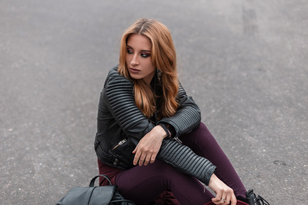 Beautiful sexy young woman in black leather jacket with a backpack in trendy pants relaxes sitting on the road in the city in the spring day. Red-haired urban girl enjoys the weekend. Retro style.の写真素材