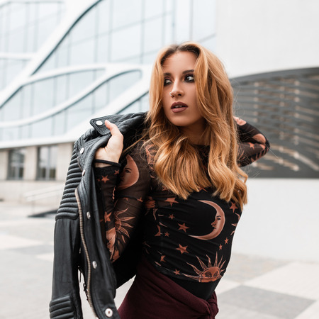 Sexy red-haired young woman with beautiful make-up with an earring in nose in a stylish black leather jacket in a vintage shirt with a pattern of moon and sun poses in a city near a modern building.の写真素材
