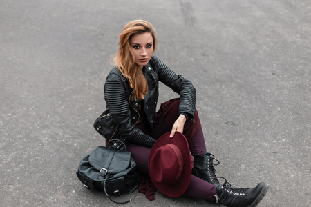Pretty young woman with a hat in a stylish leather jacket in purple leggings in black boots with a trendy backpack sits on the asphalt on the street. Beautiful red-haired girl resting outdoors.Fashionの写真素材
