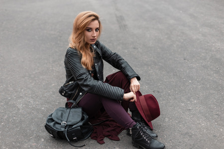 Stylish young woman with a hat in a vintage leather jacket in purple pants in black boots with a black backpack is resting sitting on a gray road outdoors. Red-haired attractive girl relaxes. Style.の写真素材