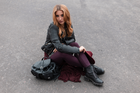 Cute pretty young woman in a stylish leather jacket in purple pants in vintage black shoes with a fashionable backpack sits on the asphalt in the city. European girl with red hair resting. Youth styleの写真素材