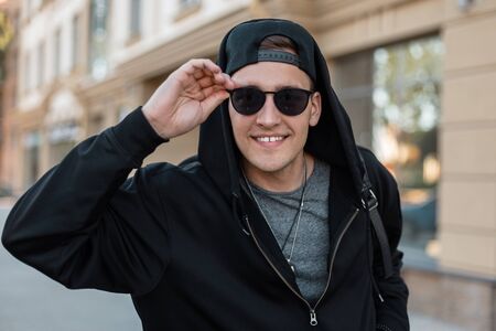 Happy handsome young hipster man in stylish sunglasses in a fashionable sweatshirt with a backpack in a cap is standing and smiling near a vintage building on the street. Joyful guy outdoors.の写真素材