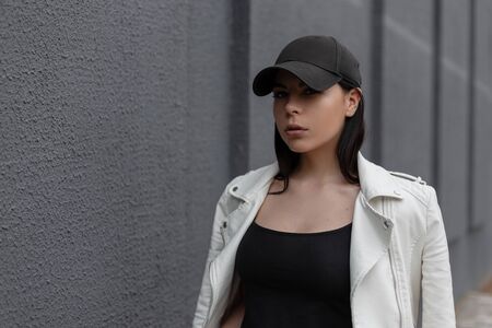 Portrait of a beautiful young brunette woman in a stylish black cap in a vintage white leather jacket in a black t-shirt near a gray wall on the street. Fashionable modern girl fashion model.  Youth.の写真素材