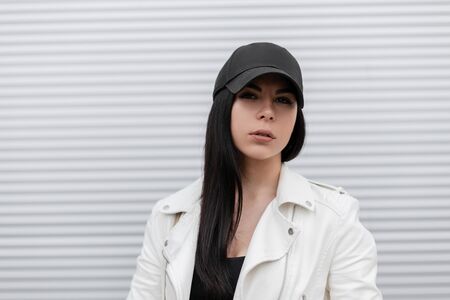 Portrait of a pretty attractive brunette young woman in a fashionable black baseball cap in a stylish white leather jacket near a modern metal wall. Beautiful attractive girl model outdoors. Fashion.の写真素材