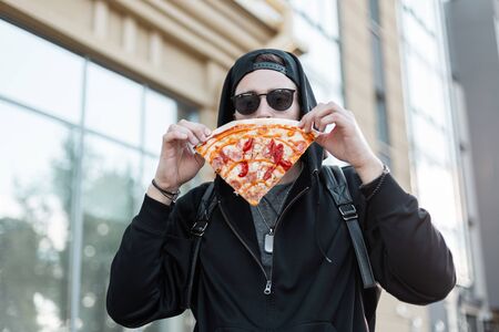 Nice young hipster male in trendy sunglasses in a black trendy hooded sweatshirt in a cap holds a slice of pizza near his face outdoors in the city. Attractive guy took a break for lunch.の写真素材