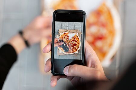 Young man makes a photo of delicious pizza on a smartphone. Closeup of male hands and pizza image on screen.  Top view.の写真素材