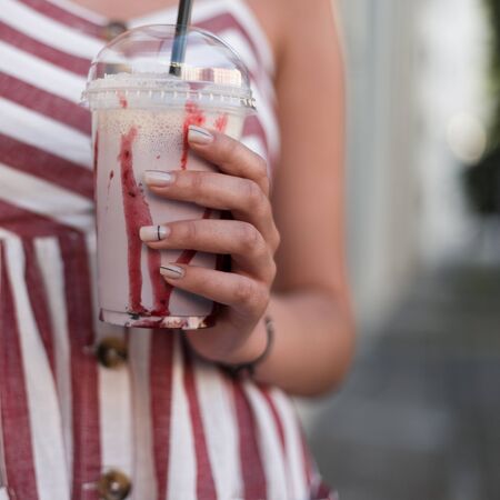 Closeup of raspberry sweet milkshake in the hands of a fashionable young woman. Stylish girl in a striped dress with a cool tasty drink on a hot summer day.の写真素材