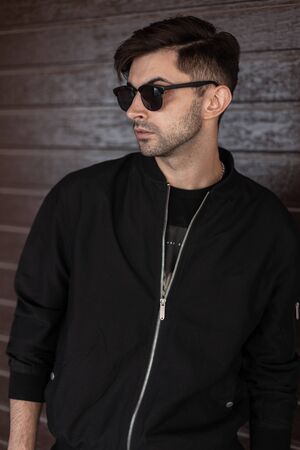 Modern handsome young man in trendy black jacket in stylish sunglasses with a hairstyle poses near a...