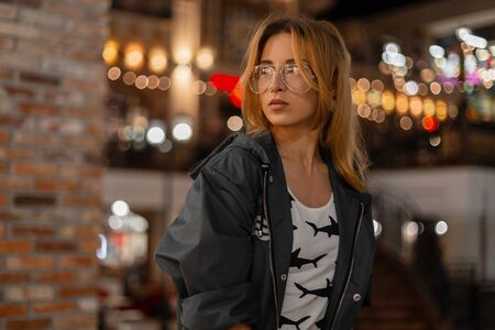 Modern pretty young woman in stylish glasses in a trendy jacket in a white t-shirt with a pattern...