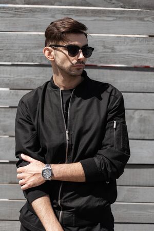 Handsome young hipster man with fashionable hairstyle in trendy black jacket in a stylish dark sunglasses poses near a gray vintage wall in a sunny summer day. Attractive brunette guy outdoors.の写真素材