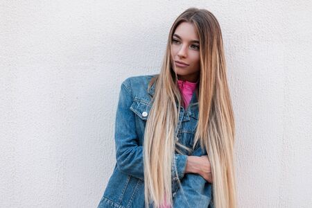 Cute young glamor woman with luxurious blond long hair in a fashionable jeans shirt in a stylish pink t-shirt poses near a vintage white wall outdoors on a summer day. Pretty attractive girl model.の写真素材