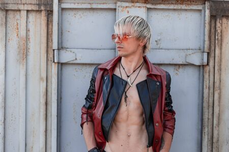 Sexy young man blond with a naked torso in a fashionable leather jacket in trendy red sunglasses is...