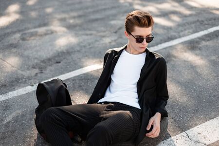 Handsome young man with trendy hairstyle relaxes on asphalt on a sunny day. Fashionable hipster guy in stylish clothes with a backpack is resting on the street and enjoying the sun. Street fashion.の写真素材
