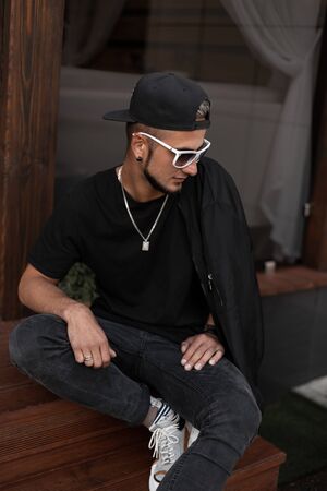 American hipster young man with a beard in a cap in a T-shirt in a jacket in sunglasses sits on wooden stairs near a vintage cafe. Handsome guy is resting in the city. Fashionable men's clothing.の写真素材
