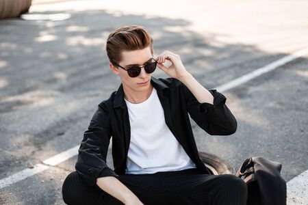 Stylish young hipster man in trendy sunglasses in an elegant shirt in striped pants resting on a sunny day outdoors. Handsome guy model enjoys sunny summer day sitting on the asphalt in the city.の写真素材