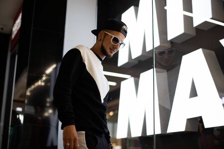 Urban cool hipster man in a stylish sweatshirt in a black fashionable black baseball cap in white sunglasses in a mall. Handsome guy model walks around the store. New collection of men's clothing.の写真素材
