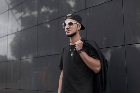 Attractive young hipster man in a fashionable jacket in stylish sunglasses with trendy black cap stand in the city on a summer day. Cool guy posing near the gray building outdoors. Street style.の写真素材