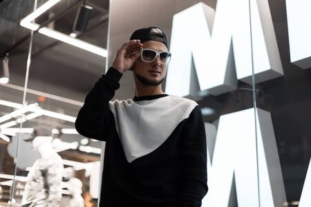 Stylish young hipster man straightens trendy sunglasses. Handsome guy with a beard in fashionable clothes in a black baseball cap posing in a modern shopping center near the glass wall. Youth style.の写真素材