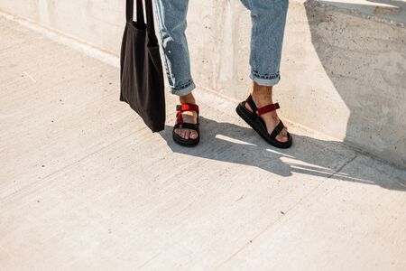 Closeup of a male legs in trendy red leather sandals in blue vintage jeans with a black fabric bag....