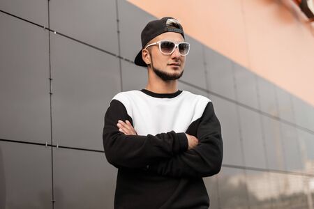 Stylish young hipster man in stylish sweatshirt in black trendy black baseball cap in white glasses...