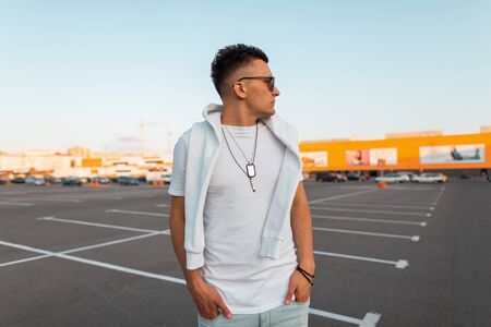 American urban young man in fashionable white summer clothes in trendy black sunglasses with a fashionable hairstyle stands on a bright sunny day in a city parking . Handsome guy hipster. Street styleの写真素材