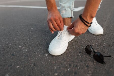 Close-up of male legs in blue vintage jeans in white fashionable leather sneakers. Trendy guy stands on the asphalt and straightens the laces on shoes.の写真素材