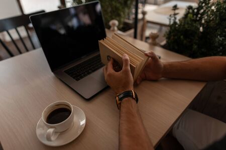 Close-up of mens hands with a book. Young man sits in a cafe with a laptop with a cup of coffee and reads an interesting book.の写真素材