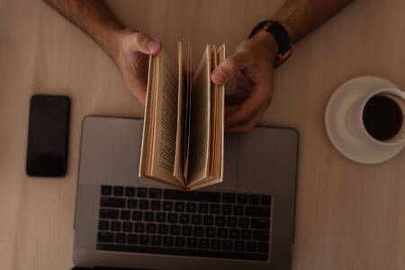 Young student man opens and reading a book while sitting at a table indoors. Top view on a wooden...