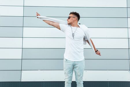 Young stylish hipster man in summer white and jeans clothes in black fashionable sunglasses poses near a modern building outdoors. American handsome guy walks around the city. Street style. Menswear.の写真素材