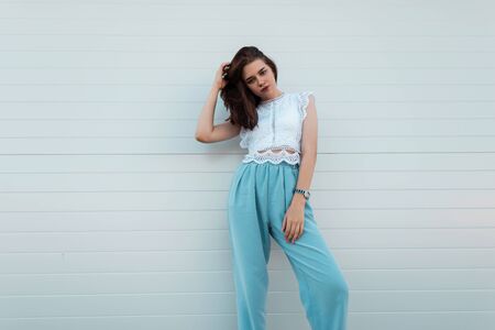 Elegant pretty young woman straightens hair. Cute girl in a trendy lace blouse in vintage blue pants posing on the street near a white wall. New summer collection of women's clothing. Stylish look.の写真素材
