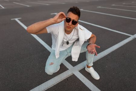 Attractive urban young man hipster in sunglasses in fashionable clothes in trendy white sneakers poses on a asphalt. Handsome urban guy sit the parking lot in the city. Stylish youth summer menswear.の写真素材