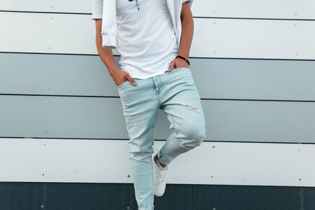 Fashionable young man in blue trendy jeans in a stylish white t-shirt in leather shoes posing near a...