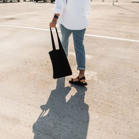 Close-up of a male body in white shirt in red leather sandals in blue jeans with a black fabric bag. Young man in trendy clothes is walks on the asphalt on a sunny day. Trendy casual summer look.の写真素材