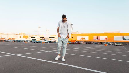 Young modern trendy hipster man in stylish dark sunglasses in vintage blue jeans in a fashionable white t-shirt in sneakers walks on the city. Handsome guy model rests outdoors. Summer street style.の写真素材