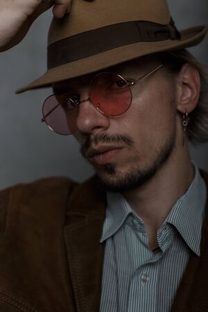 Close-up face of brutal young hipster man in trendy round glasses in elegant retro clothes in vintage hat in the studio near gray wall. Bearded sexy guy indoors. Old fashioned style. Elegant menswear.の写真素材