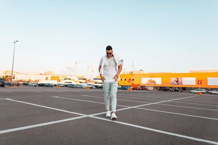 Young modern trendy hipster man in stylish dark sunglasses in vintage blue jeans in a fashionable white t-shirt in sneakers walks on the city. Handsome guy model rests outdoors. Summer street style.の写真素材