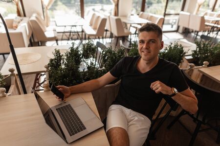 Happy young man in a fashionable black t-shirt is resting while sitting at a table in a vintage cafe with a modern laptop and with a phone in hands. Successful joyful freelancer guy relaxes indoors.の写真素材
