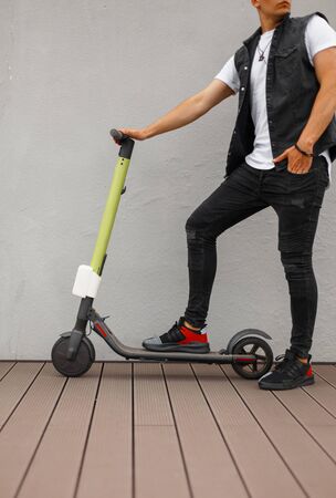 Stylish young man in gray jeans in a denim vest in a T-shirt in fashionable sneakers stands on a modern scooter near the wall. Trendy guy with electric scooter indoors. Leisure.の写真素材
