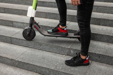 Stylish guy in trendy jeans in sneakers stands with a scooter on the stone vintage steps in the street on a summer day. Closeup of man legs on a modern electric scooter. Healthy lifestyle.の写真素材